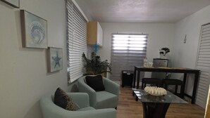 Una sala de estar con un sillón azul claro, una mesa de centro de vidrio, y un comedor con una mesa y sillas negras.