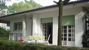 Un bâtiment blanc avec des volets verts, une table recouverte d’une nappe jaune et une chaise à l’extérieur.