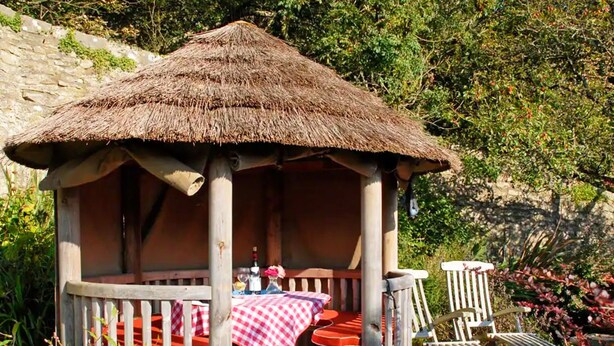 A wooden gazebo with a thatched roof, a table with a red and white checkered tablecloth, and two chairs. - Slade Cottage (Bridgend)