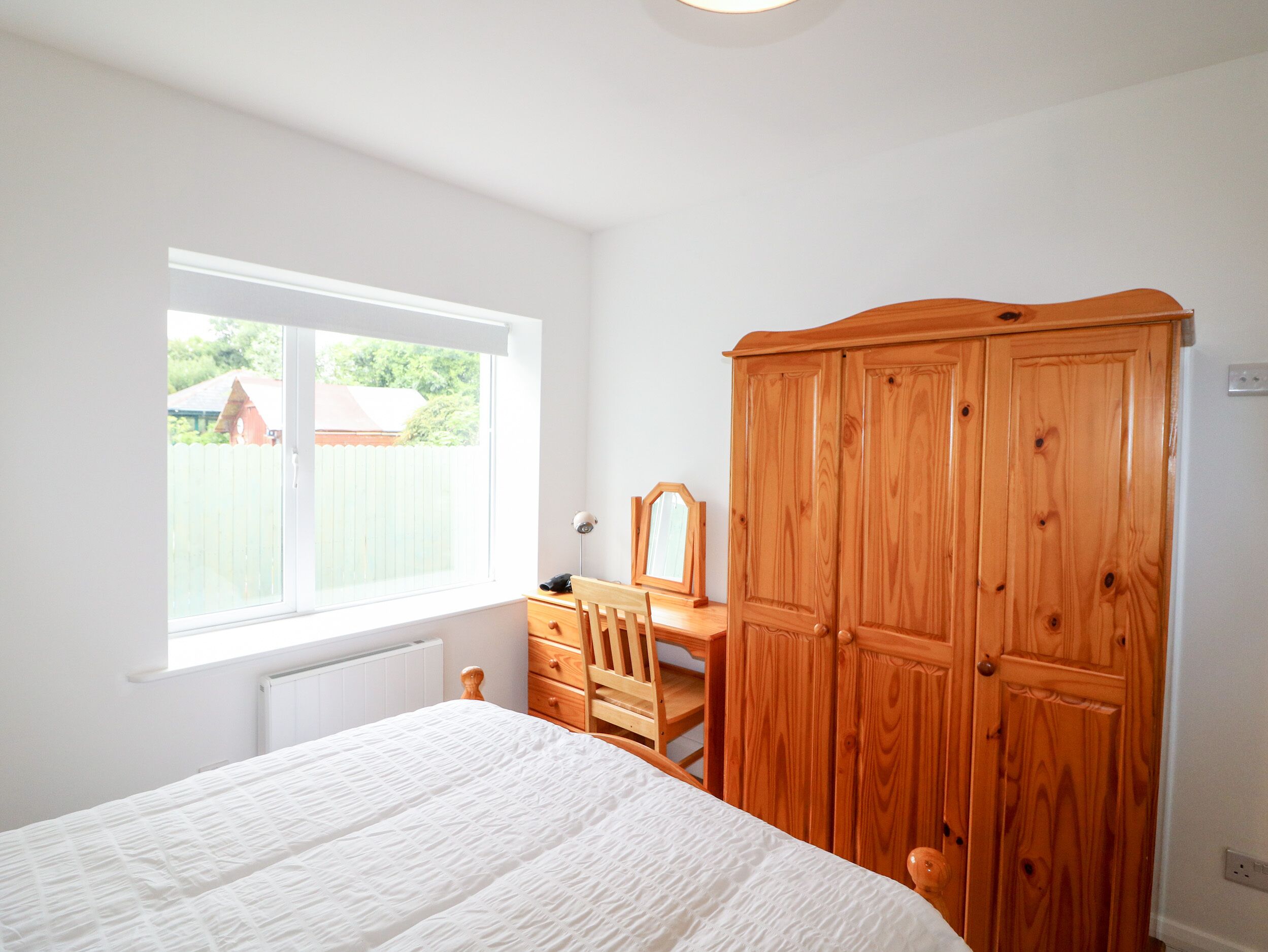 A bedroom with a bed, a wooden wardrobe, a desk with a chair, and a window with a view of a house.