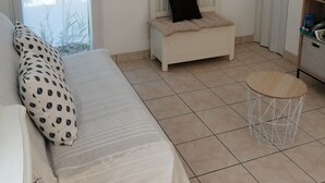 A room with a bed, a small white bench with a black cushion, a round wooden table, a framed picture on the wall, and a television on a stand. - Maison Proximitée Plage de Banastère 2 Personnes (Sarzeau)