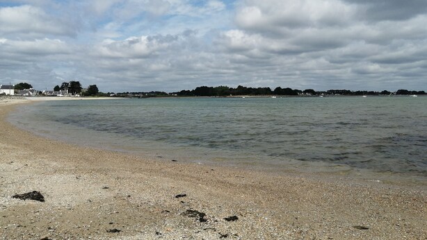 A beach with pebbly shore, calm water, and a distant shoreline with buildings and trees under a cloudy sky. - Maison Proximitée Plage de Banastère 2 Personnes (Sarzeau)