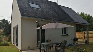 A single-story house with a patio area featuring a table and chairs, a covered seating area, and a small window. - Maison Proximitée Plage de Banastère 2 Personnes (Sarzeau)