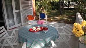 Um pátio com toalha de mesa xadrez, cadeiras brancas, uma mesa com uma bandeja de comida e uma cesta de frutas.