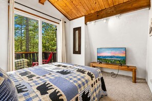 A bedroom with a large bed, a television on a wooden stand, and a view of trees through a sliding glass door. - Bear PAC Hideout (Big Bear Lake)