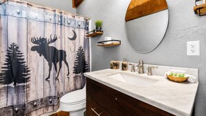 Un baño con temática rústica, que incluye una cortina de ducha con diseño de alce y bosque, un inodoro blanco, un lavamanos de mármol con espejo y armarios de madera.