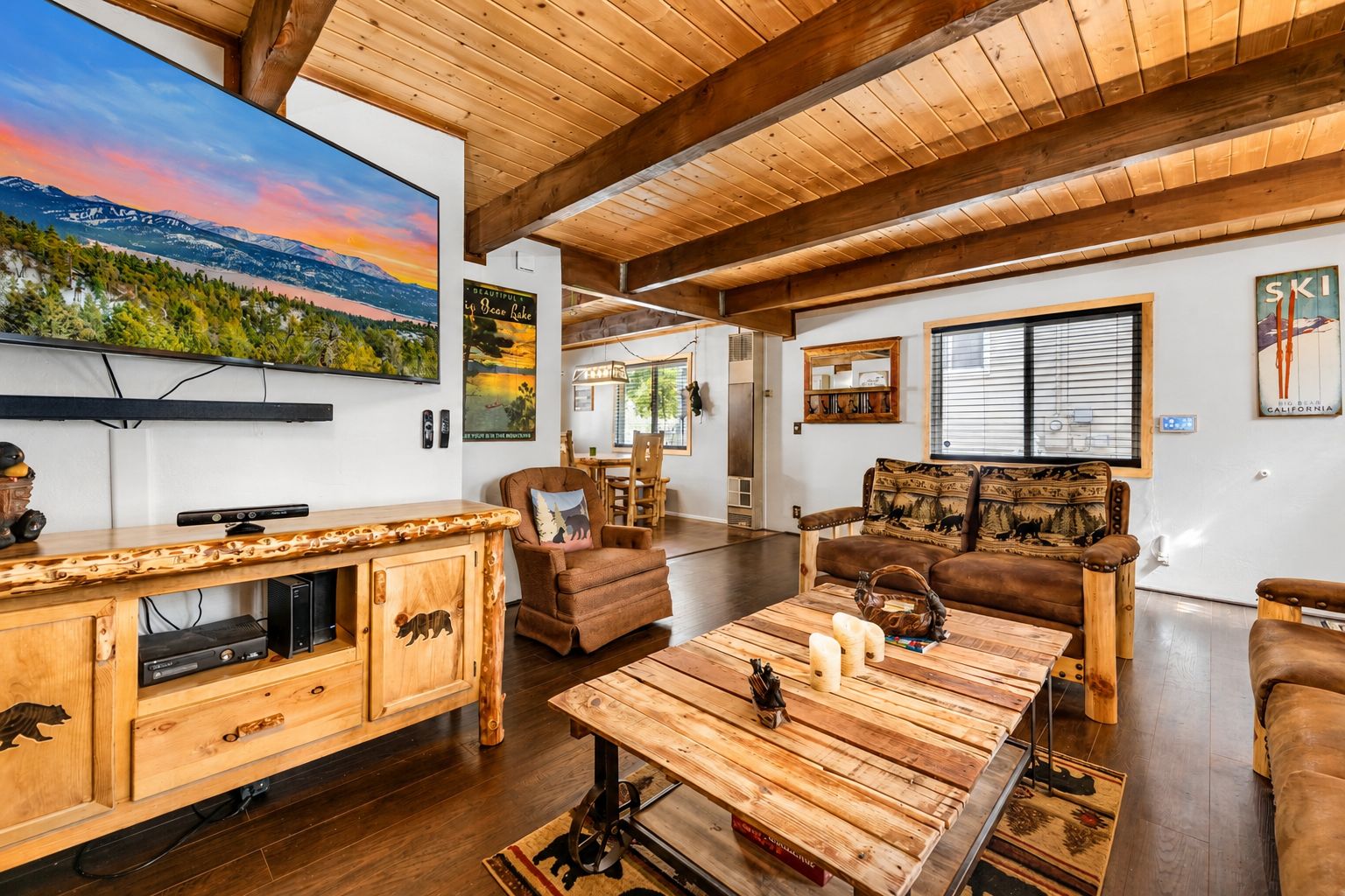 A spacious living room with a wooden ceiling, a large flat-screen TV mounted on the wall, a wooden entertainment unit, a brown leather sofa, and a coffee table with decorative items.