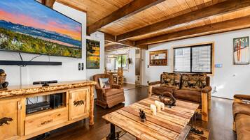 A spacious living room with a wooden ceiling, a large flat-screen TV mounted on the wall, a wooden entertainment unit, a brown leather sofa, and a coffee table with decorative items.