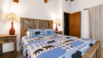 Une chambre à coucher avec une tête de lit en bois, une couvre-lit à carreaux ornée de motifs animaliers, deux tables de chevet avec des lampes et une porte en bois.