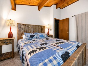 A bedroom with a wooden headboard, a checkered bedspread with animal prints, two bedside tables with lamps, and a wooden door.