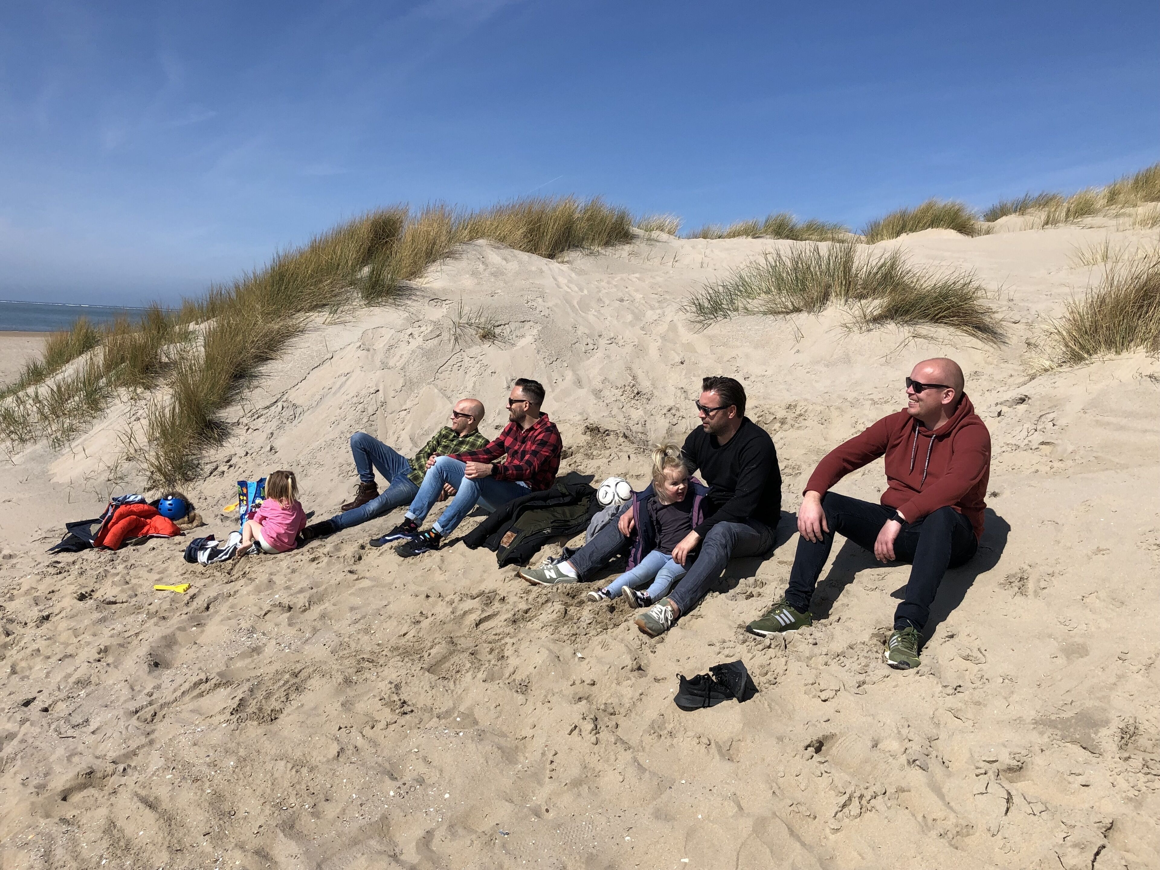 Eine Gruppe von Menschen sitzt am sandigen Strand, im Hintergrund Dünen.