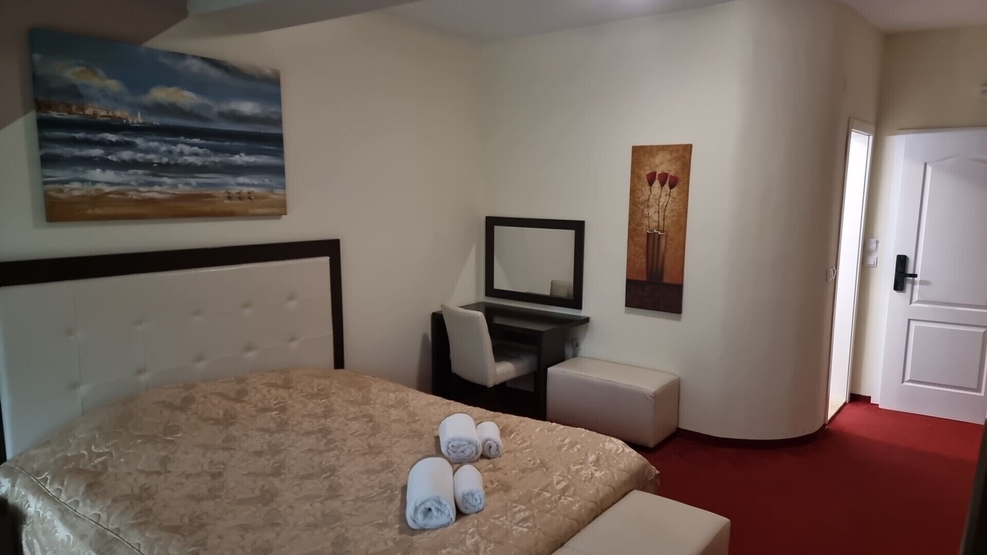 A hotel room with a bed, a desk with a chair, a small stool, a mirror, and artwork on the walls.