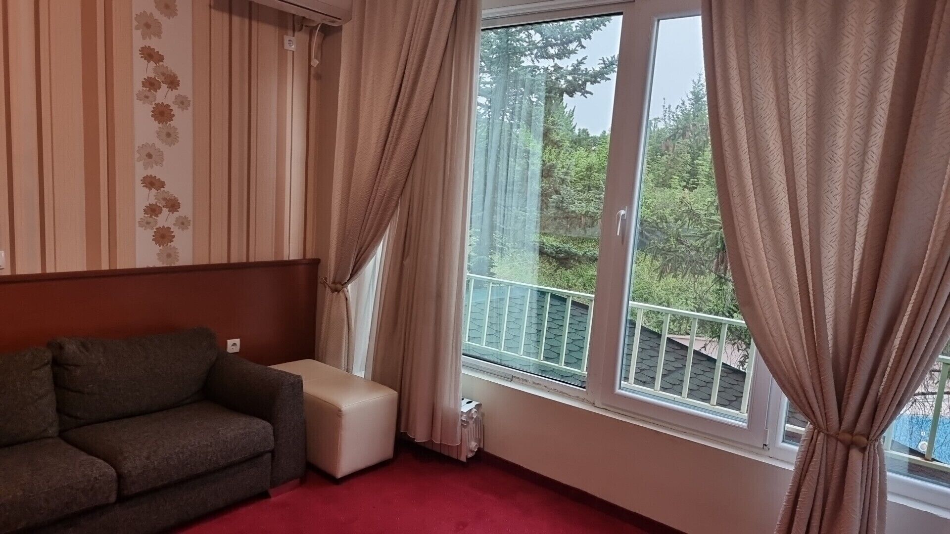 Een kamer met een bank, een klein voetenbankje, een radiator en een raam met gordijnen.