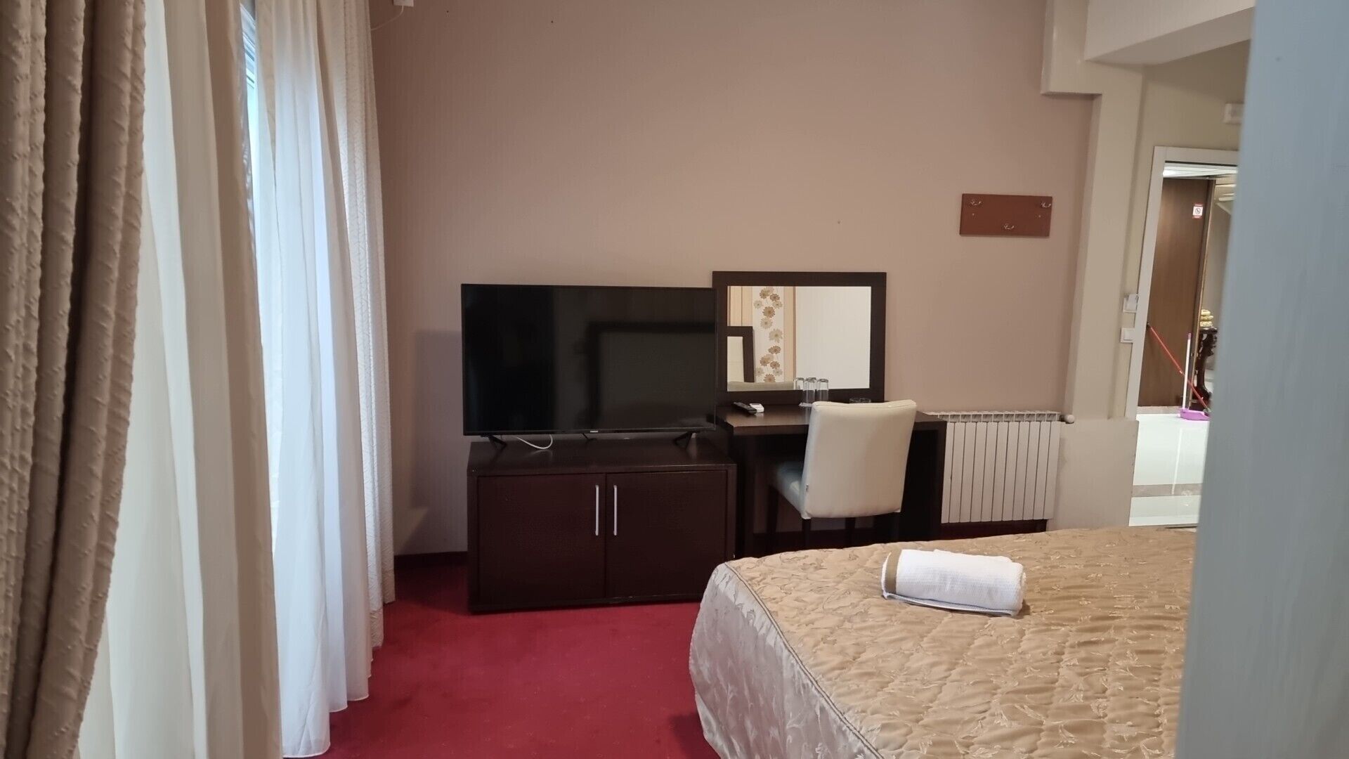 Habitación de hotel con cama, escritorio, silla, televisor, espejo y radiador.