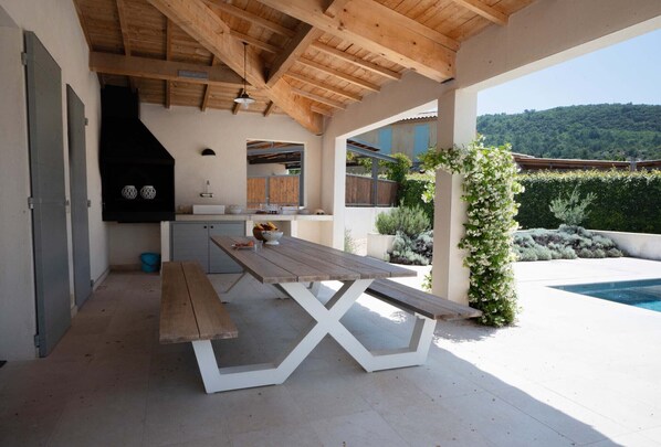 A covered outdoor kitchen area with a wooden table, a bench, and a view of a swimming pool and greenery.