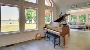 A spacious living room with a grand piano, large windows offering a view of a grassy area and a body of water, and a cozy seating area with a sofa and a coffee table. - Lakefront Sunburg Vacation Rental w/ Boat Dock! (Sunburg)