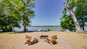 A beachside patio with Adirondack chairs and a fire pit, overlooking a lake with a dock and boat. - Lakefront Sunburg Vacation Rental w/ Boat Dock! (Sunburg)
