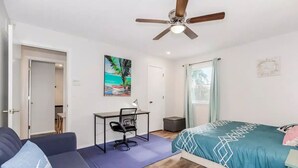 Ein Schlafzimmer mit Bett, Schreibtisch, Stuhl, Deckenventilator und einer Tür zu einem weiteren Raum.