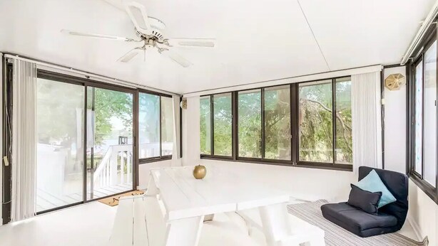 Ein geräumiges Zimmer mit einem weißen Tisch, einem Deckenventilator, großen Fenstern und einem schwarzen Sofa mit Kissen.