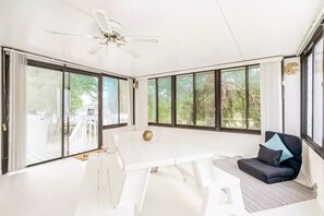 Una habitación amplia con una mesa blanca, un ventilador de techo, ventanas grandes y un sofá negro con cojines.
