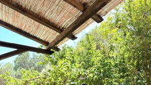 Una mesa de madera con una botella de vino y dos copas, colocada al aire libre bajo una pérgola de madera y con árboles verdes exuberantes al fondo.