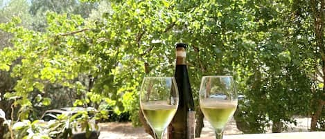 A wooden table with a bottle of wine and two glasses on it, set outdoors with a wooden pergola overhead and lush green trees in the background.
