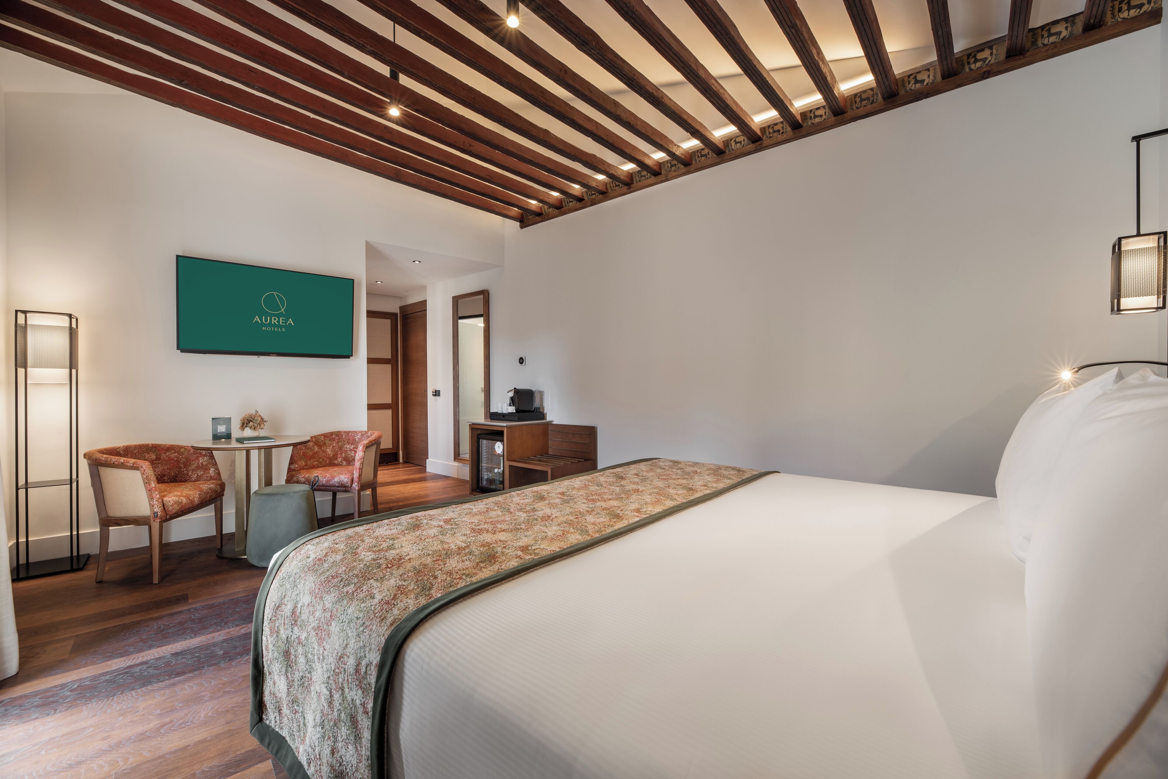 Una habitación de hotel con una cama, dos sillones tapizados, una mesita y un televisor montado en la pared.