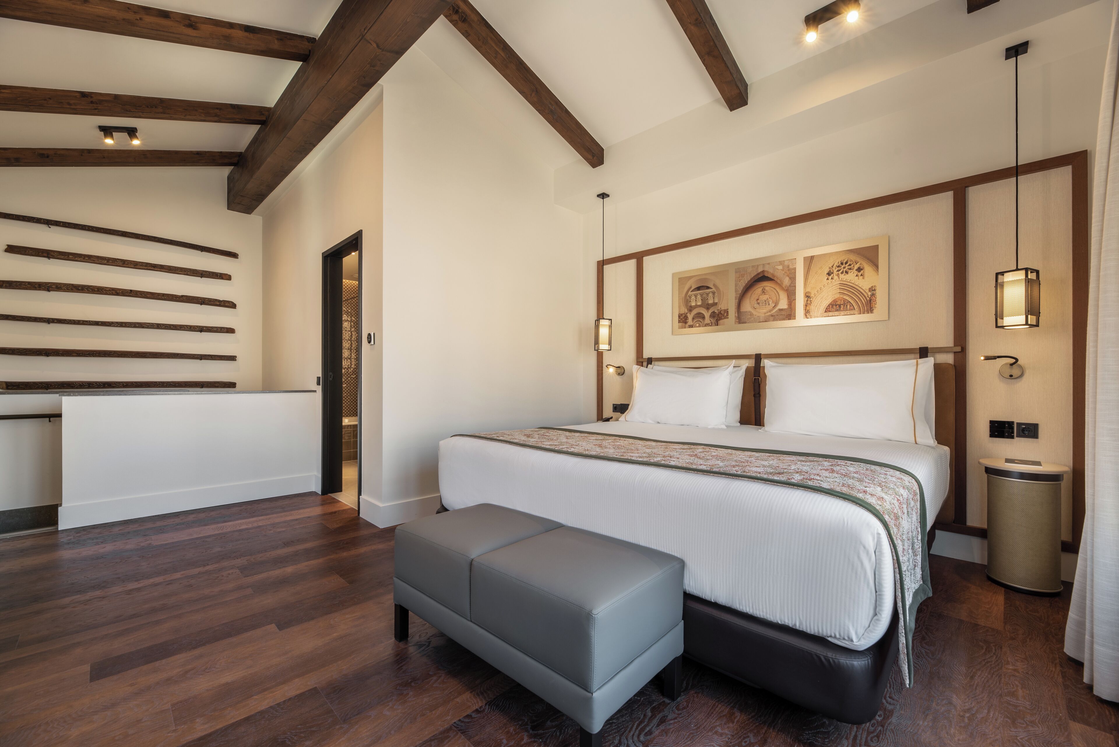 A modern bedroom with a large bed, a bench, wooden beams, and artwork on the wall.