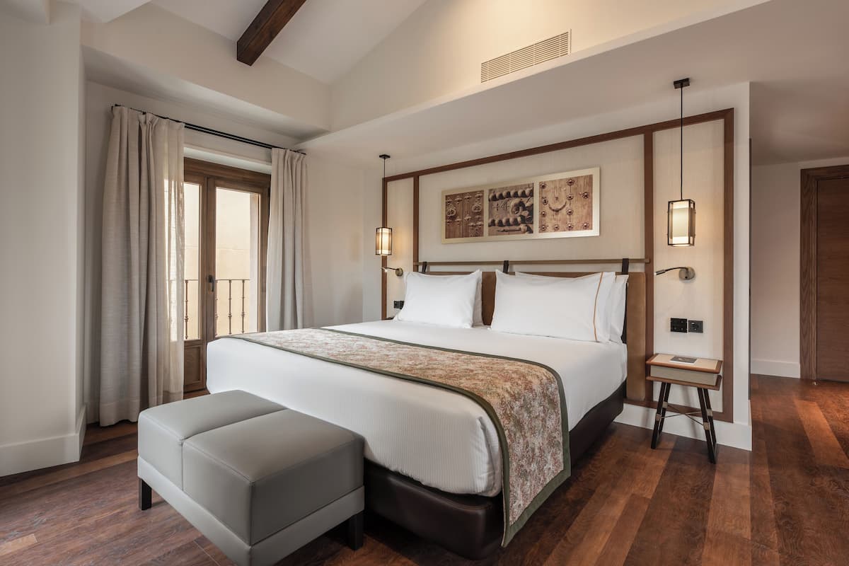 Habitación de hotel con una cama grande, un banco, una mesita pequeña y una ventana con cortinas.