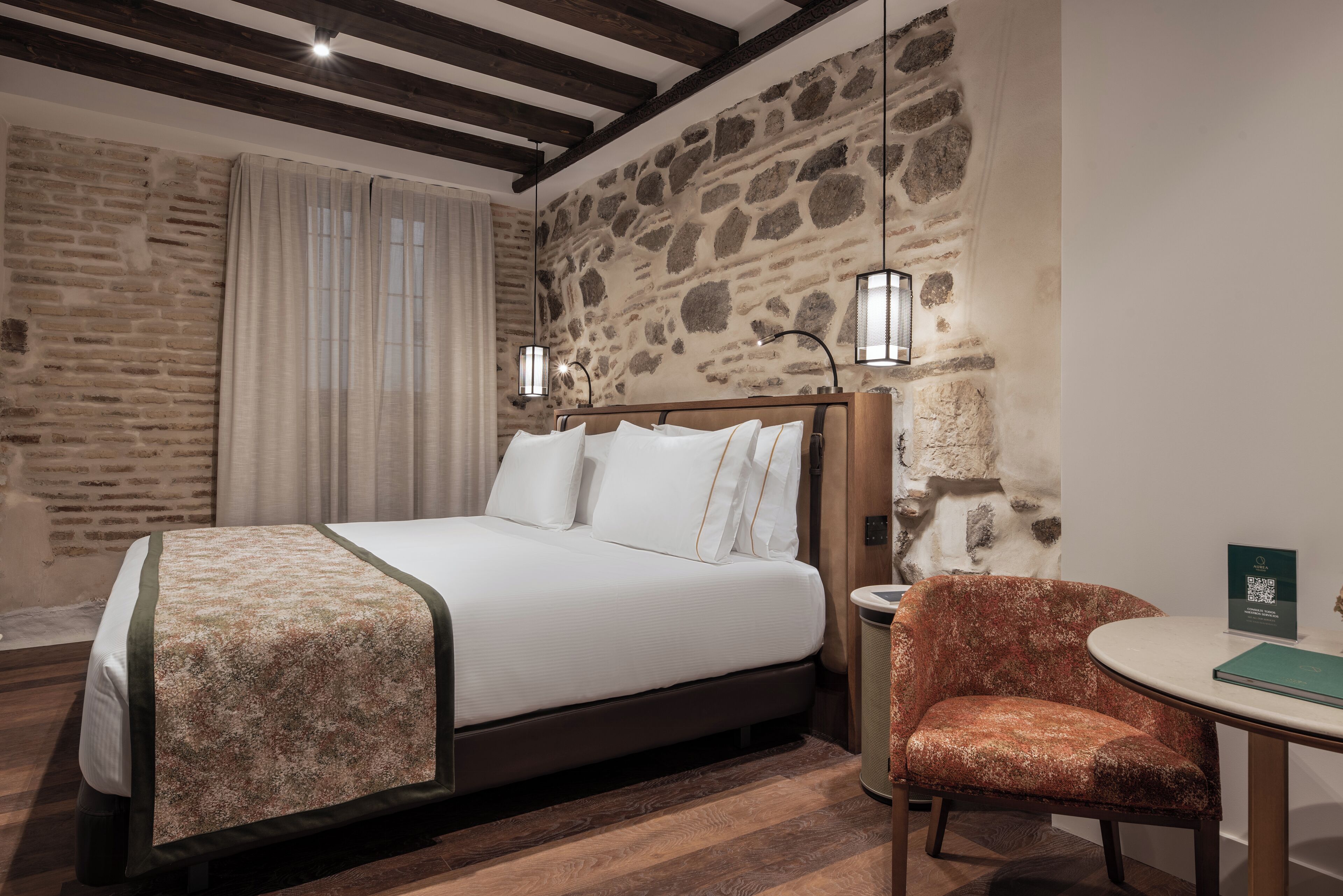 Una habitación de hotel con una cama grande, una silla, una mesita y paredes de piedra.