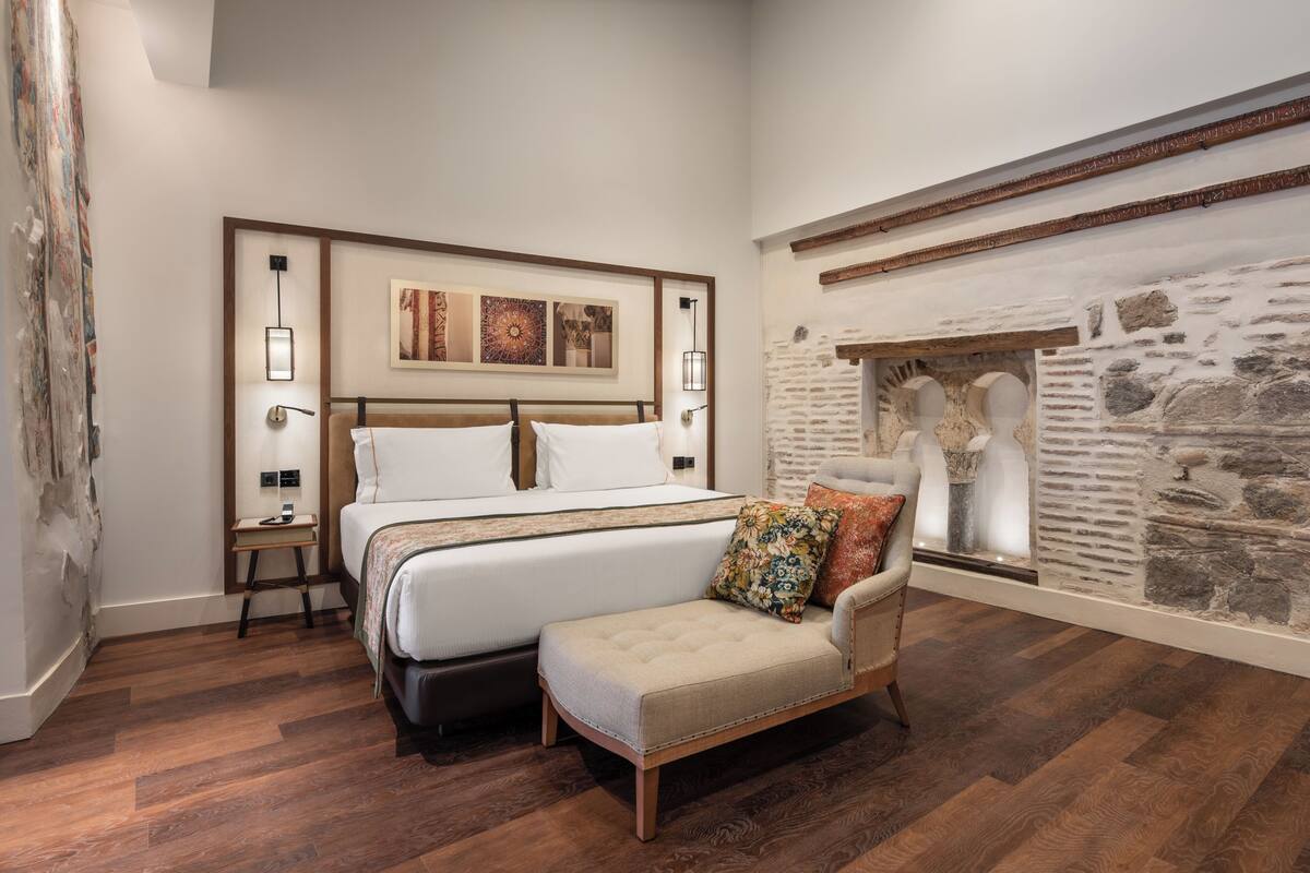 Un dormitorio con una cama grande, una mesita de noche, una silla y una pared de piedra.
