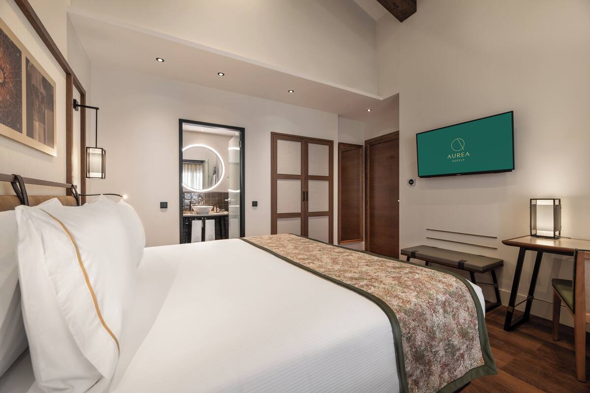 Habitación de hotel con una cama grande, un escritorio, una banca, un televisor y un baño visible a través de una puerta abierta.