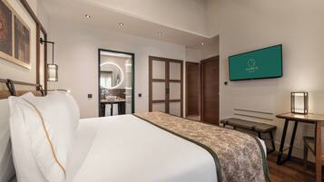 A hotel room with a large bed, a desk, a bench, a TV, and a bathroom visible through an open door.