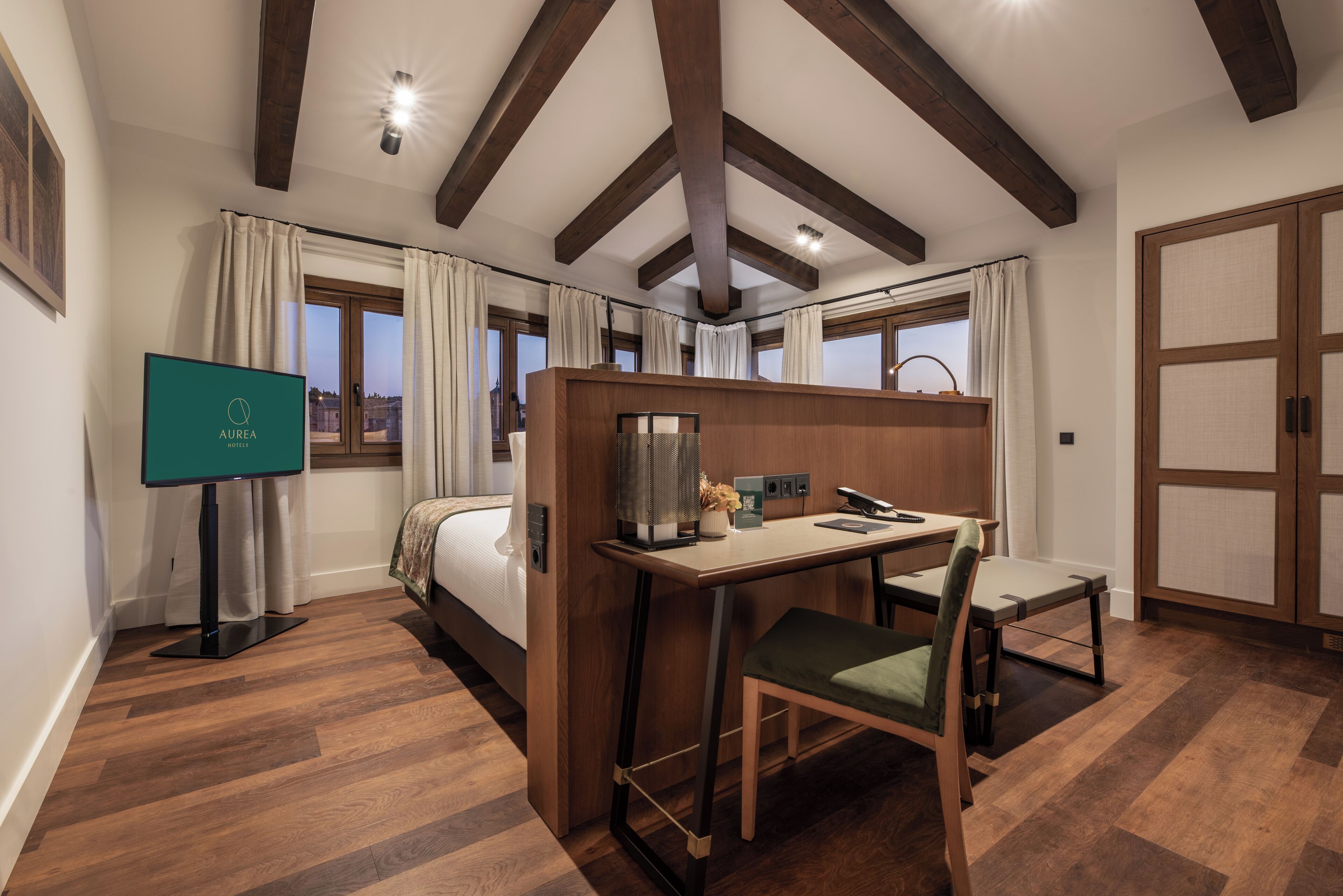 A hotel room with a bed, a desk with a chair, a television, and a view of the city through large windows.