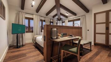 A hotel room with a bed, a desk with a chair, a television, and a view of the city through large windows.