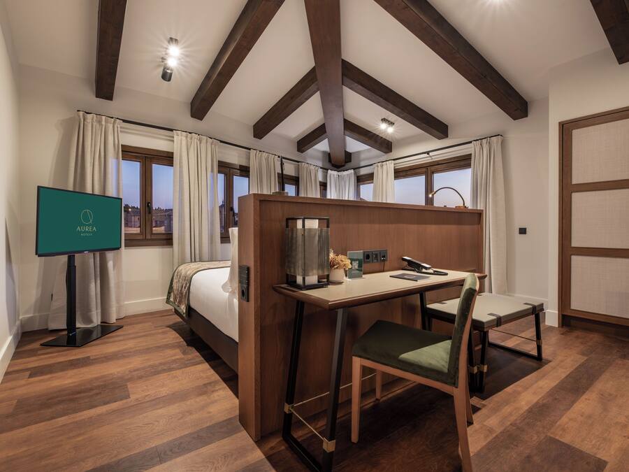 A hotel room with a bed, a desk with a chair, a television, and a view of the city through large windows.