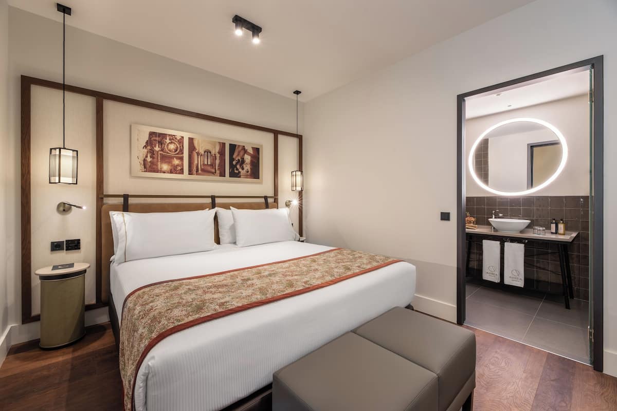 Habitación de hotel con una cama grande, lámparas de noche, cabecero con obra de arte, espejo redondo con luces, y baño con lavamanos y toalleros.