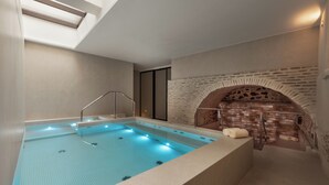 A modern indoor swimming pool with a clear blue water surface, a built-in bench, and a brick wall with a fireplace. - Áurea Toledo (Toledo)