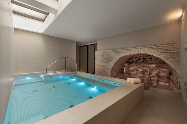 Una piscina interior moderna con superficie de agua azul cristalina, una banca integrada y una pared de ladrillo con chimenea.