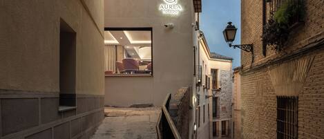 Une ruelle étroite, dont on aperçoit l’intérieur moderne à travers une fenêtre, une enseigne indiquant « Aurea Toledo » et des bâtiments traditionnels en briques.