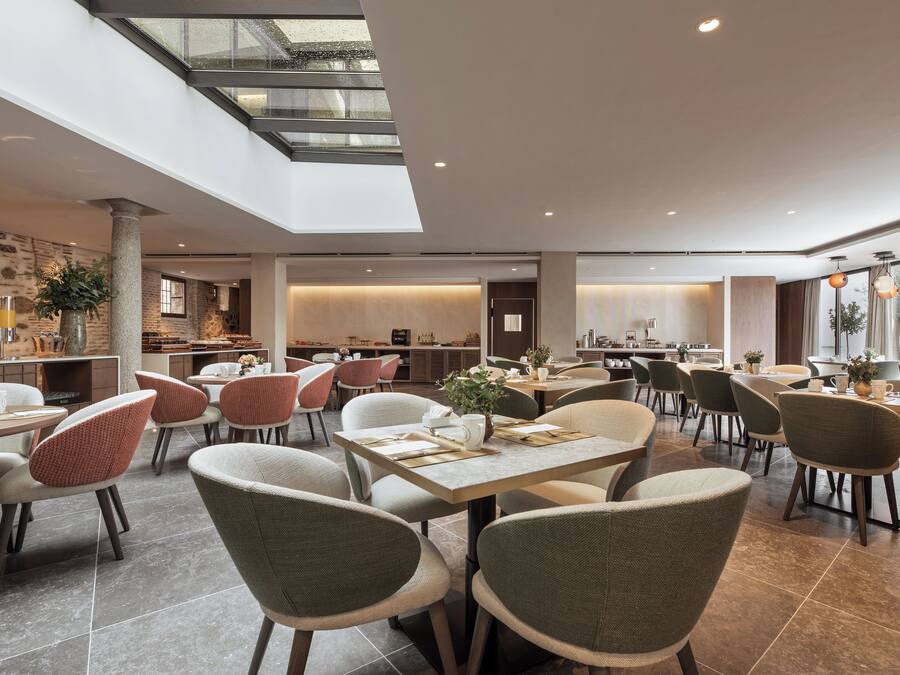 A spacious dining area with multiple tables and chairs, a high ceiling with skylights, and a modern kitchen in the background.