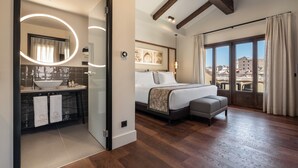 A modern bedroom with a large bed, a bedside table, a small sofa, a bathroom with a sink and mirror, and a view of buildings through the window. - Áurea Toledo (Toledo)