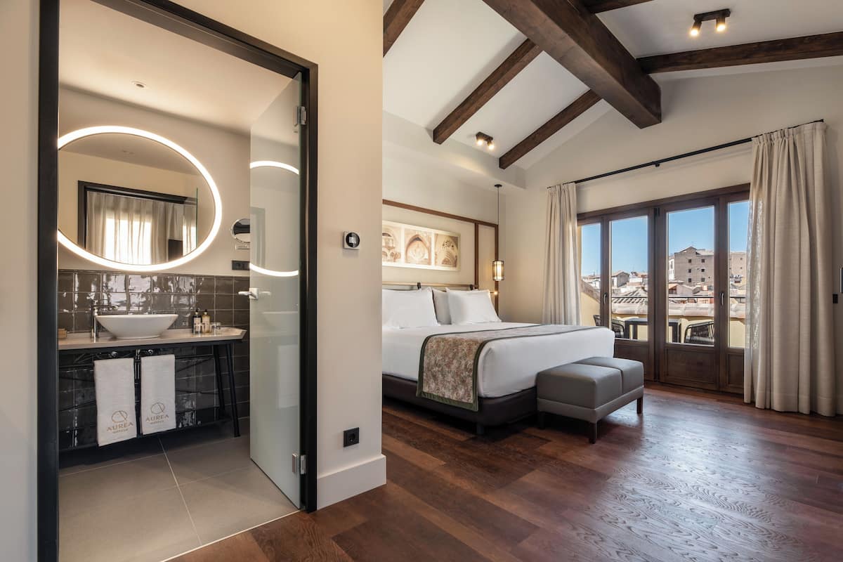 Un dormitorio moderno con una cama grande, una mesita de noche, un pequeño sofá, un baño con lavamanos y espejo, y vistas a edificios a través de la ventana.
