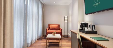 A hotel room with a sofa, a coffee table, a desk with a chair, a flat-screen TV, and a lamp.