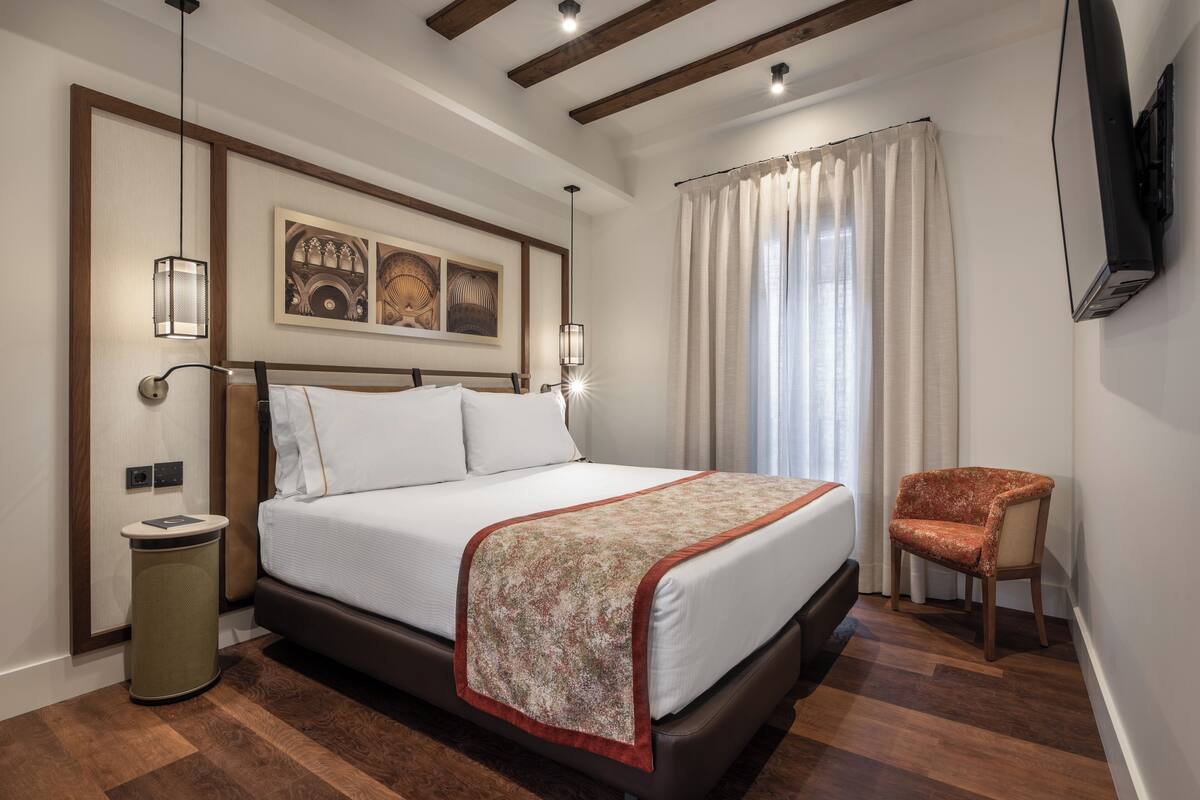 Un dormitorio ordenado con cama, una silla, un cesto de basura y un televisor instalado en la pared.