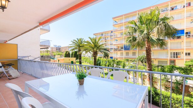 A balcony with a white table and chairs, a potted plant, and a view of a multi-story building with balconies and palm trees. - ADRIAN Apartment (La Pineda)