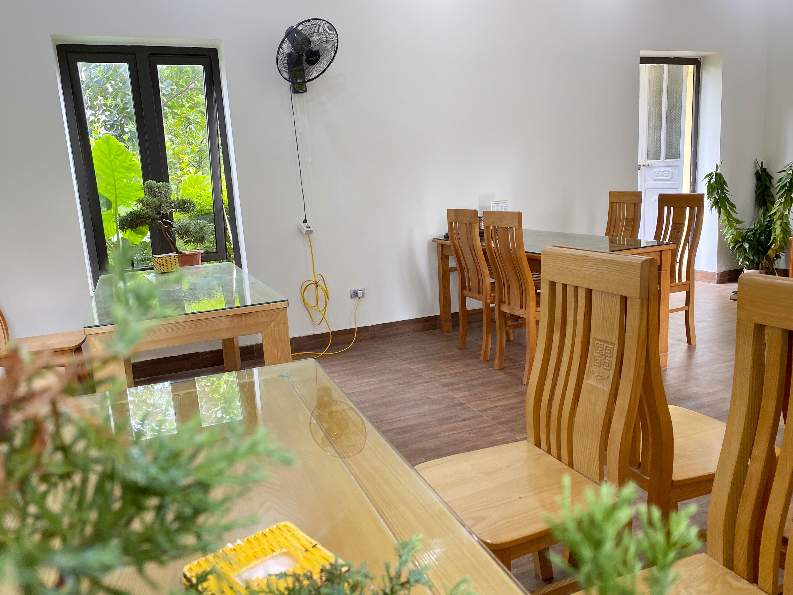 A dining area with wooden chairs and a glass table, a wall-mounted fan, and a view of greenery outside.