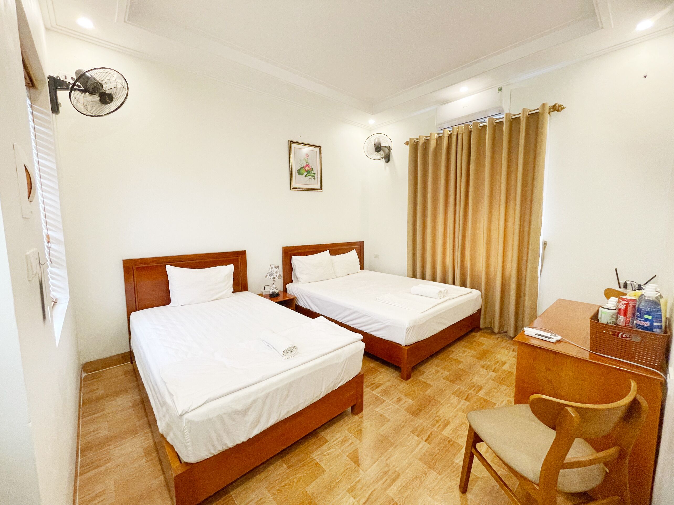Habitación de hotel con dos camas individuales, un escritorio con silla, una mesita con artículos de aseo y un ventilador de pared.