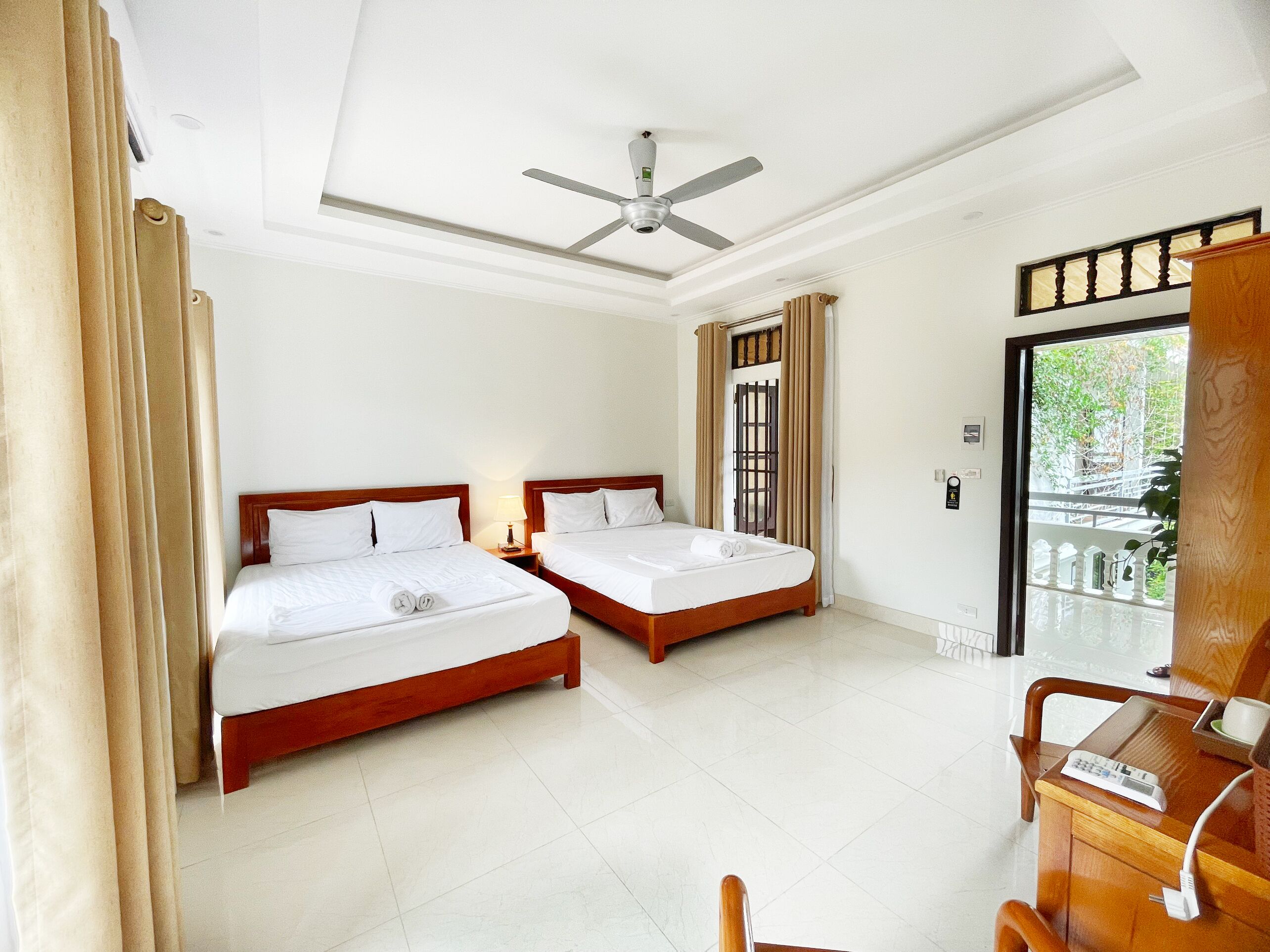 A hotel room with two beds, a ceiling fan, a wooden desk with a telephone, and a view of greenery outside.