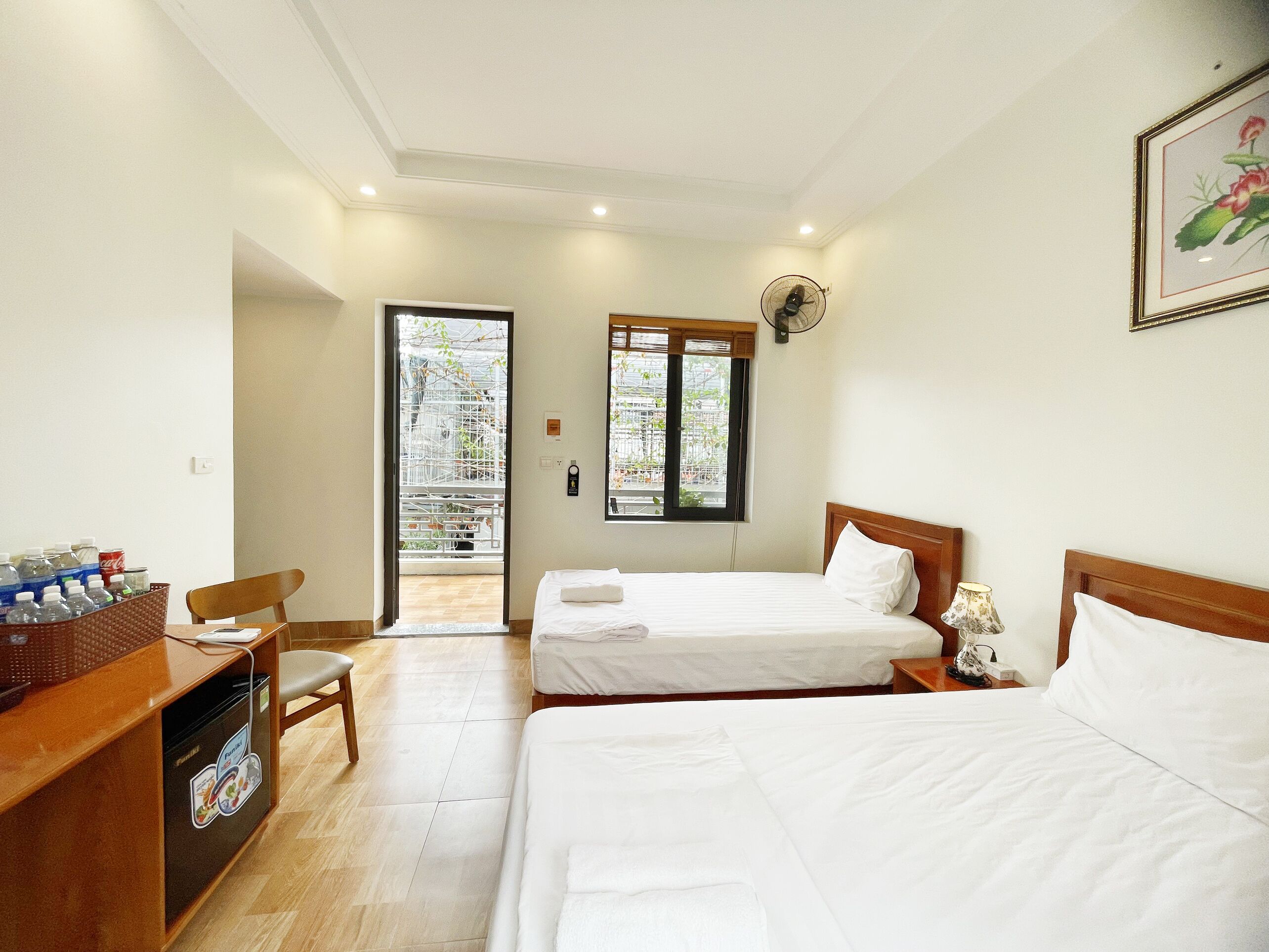 A hotel room with two beds, a desk with a chair, a fan, and a window with a view of the outside.